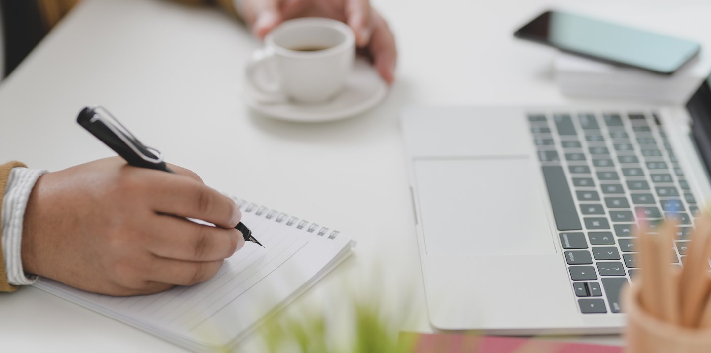 Crop journalist writing in notebook while working on article at desk with laptop and coffee